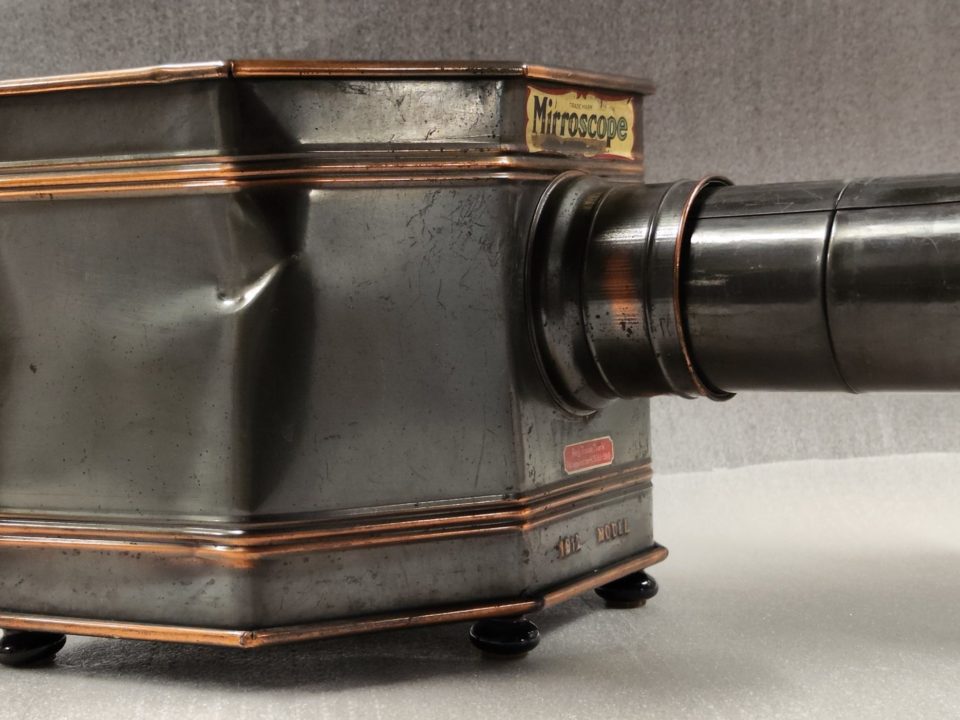 Imagen de un "Mirroscope", también conocido como linterna mágica. Es un aparato antiguo similar a una mezcla entre un proyector y una cámara reflex, fabricado en metal oscuro.